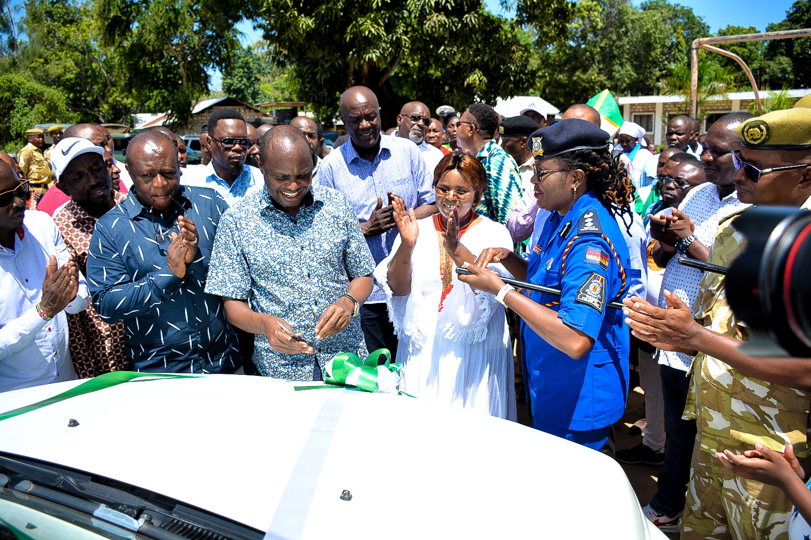 Chief of Staff and Head of Public Service, Felix Koskei (front row with blue short sleeved flowery shirt), cuts the ribbon for the donated patrol vehicle, as the Cabinet Secretary for Tourism, Wildlife and Heritage, Hon. Peninah Malonza, claps on (front row with white dress)