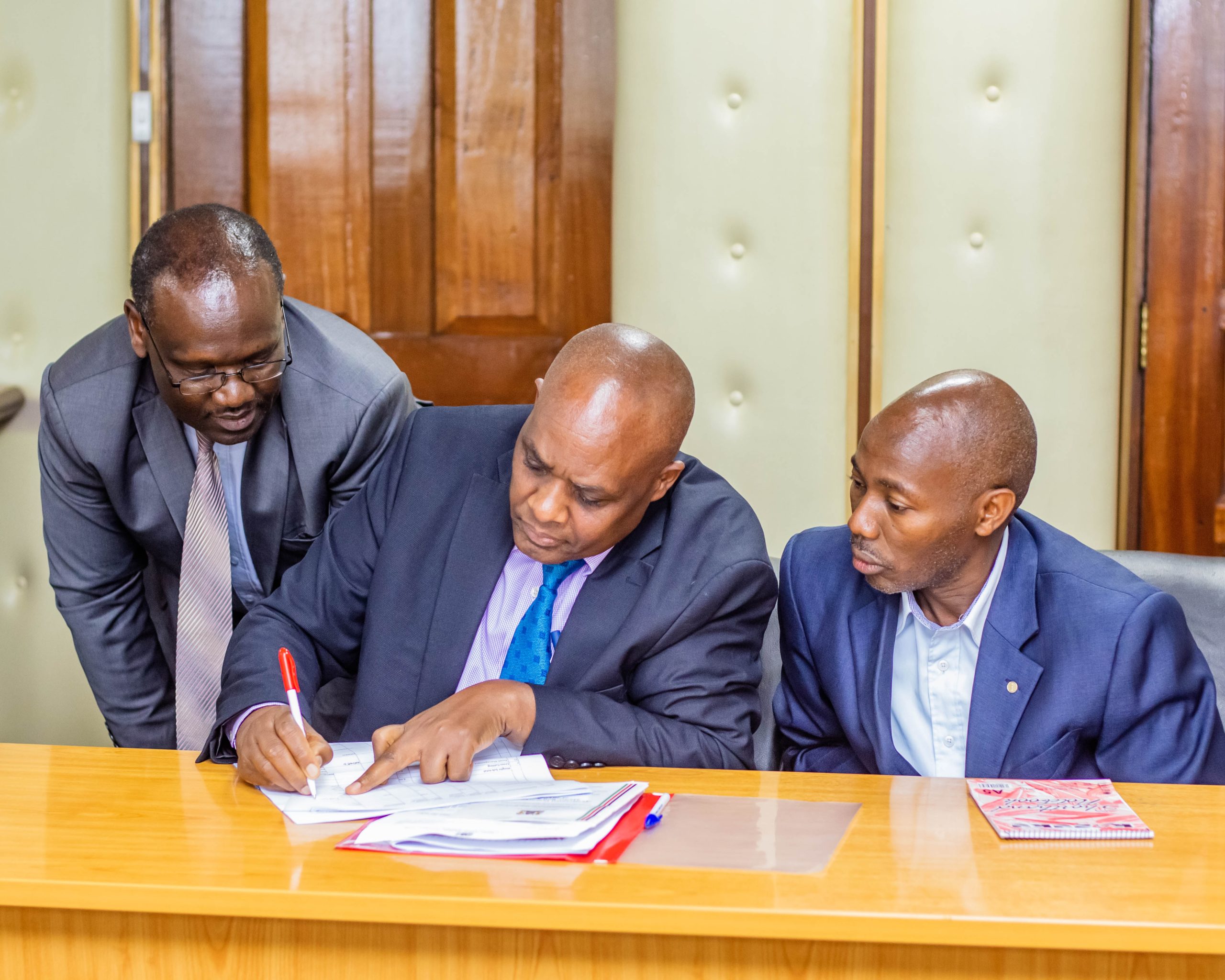 Head of Supply Chain Management Mr Justus Areri signs his Deparment's Performance Contract flanked by Head of ICT Mr Francis Muriuki [right) and Head of Planning, Charles Ombuki (Left)