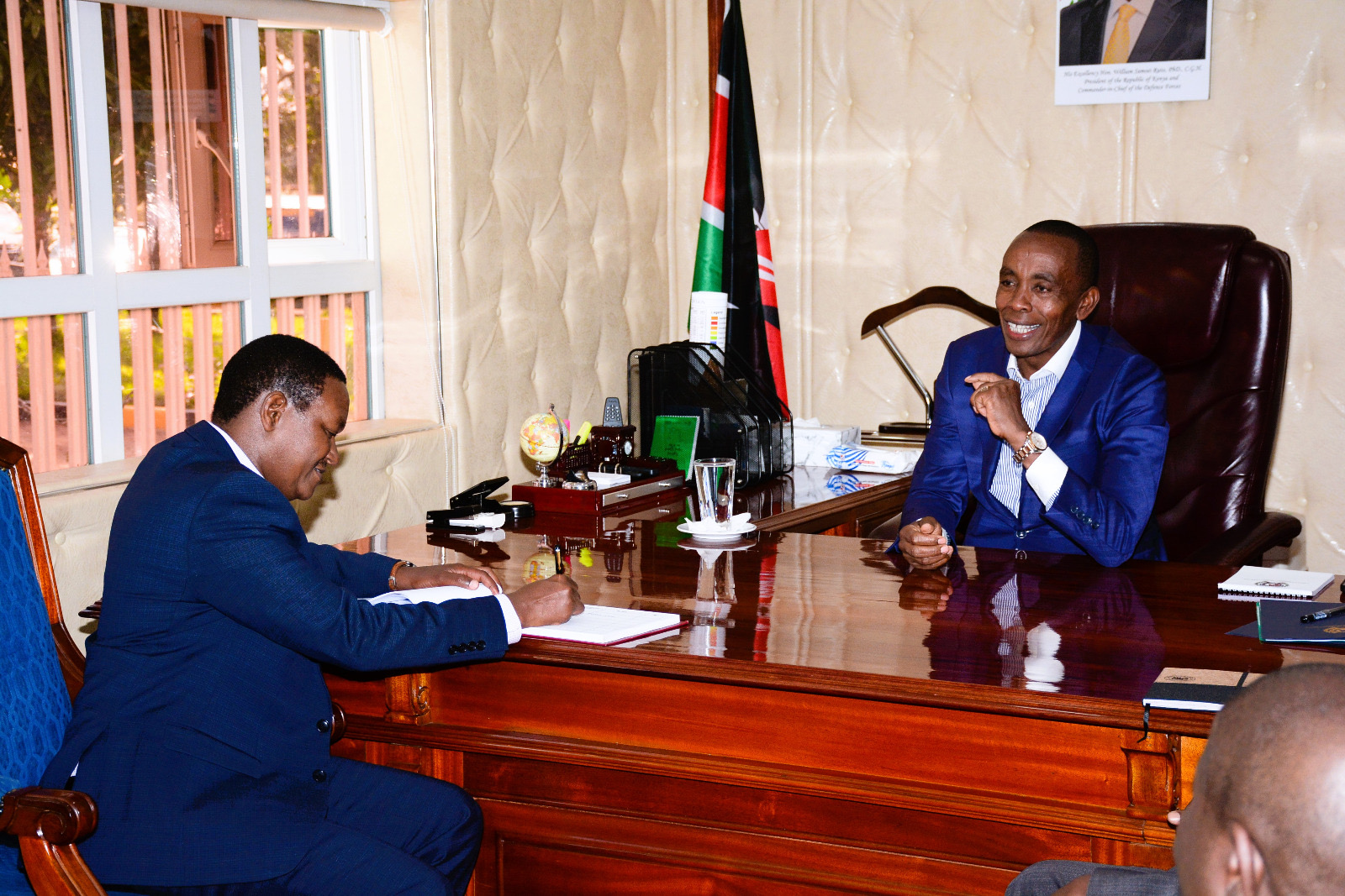 The Cabinet Secretary, Ministry of Tourism and Wildlife, Dr. Alfred Mutua (left), signs the Governor's visitors' book, as the Kiambu County Governor, H.E. Dr. Paul Wamatangi (right) looks on.